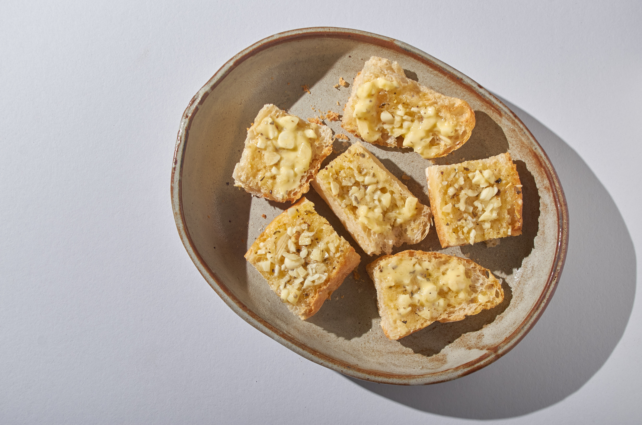 A ceramic plate with six pieces of sliced bread topped with melted cheese and minced garlic. The plate is placed on a light surface