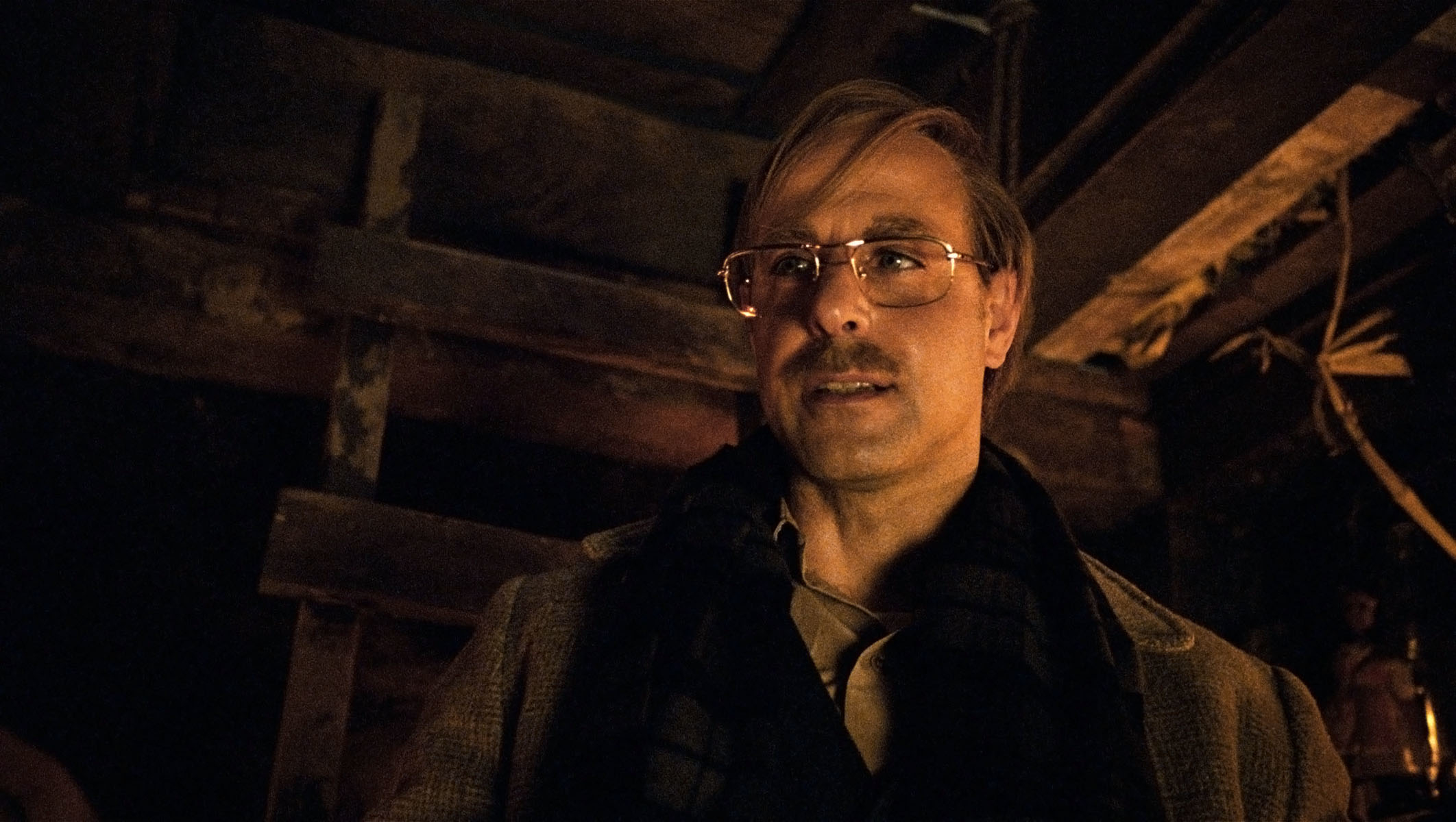 A person wearing glasses and a dark coat in a dimly lit room. They have a serious expression and are slightly looking to the side
