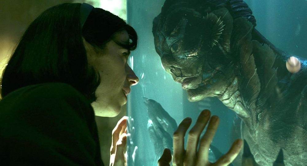 A woman with a headband and a humanoid fish creature have an intimate moment, touching their hands to the glass between them