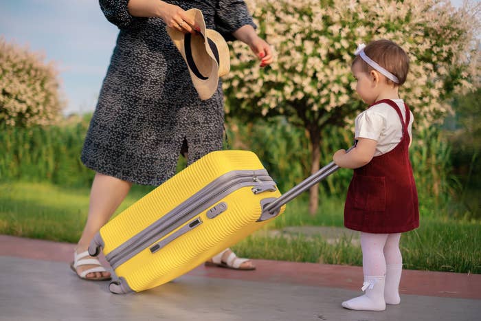 A woman holding a hat and a child pulling a yellow suitcase outdoors