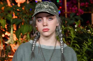 Billie Eilish wears a camouflage cap and gray sweatshirt, with braided hair. She stands in front of a backdrop of green plants