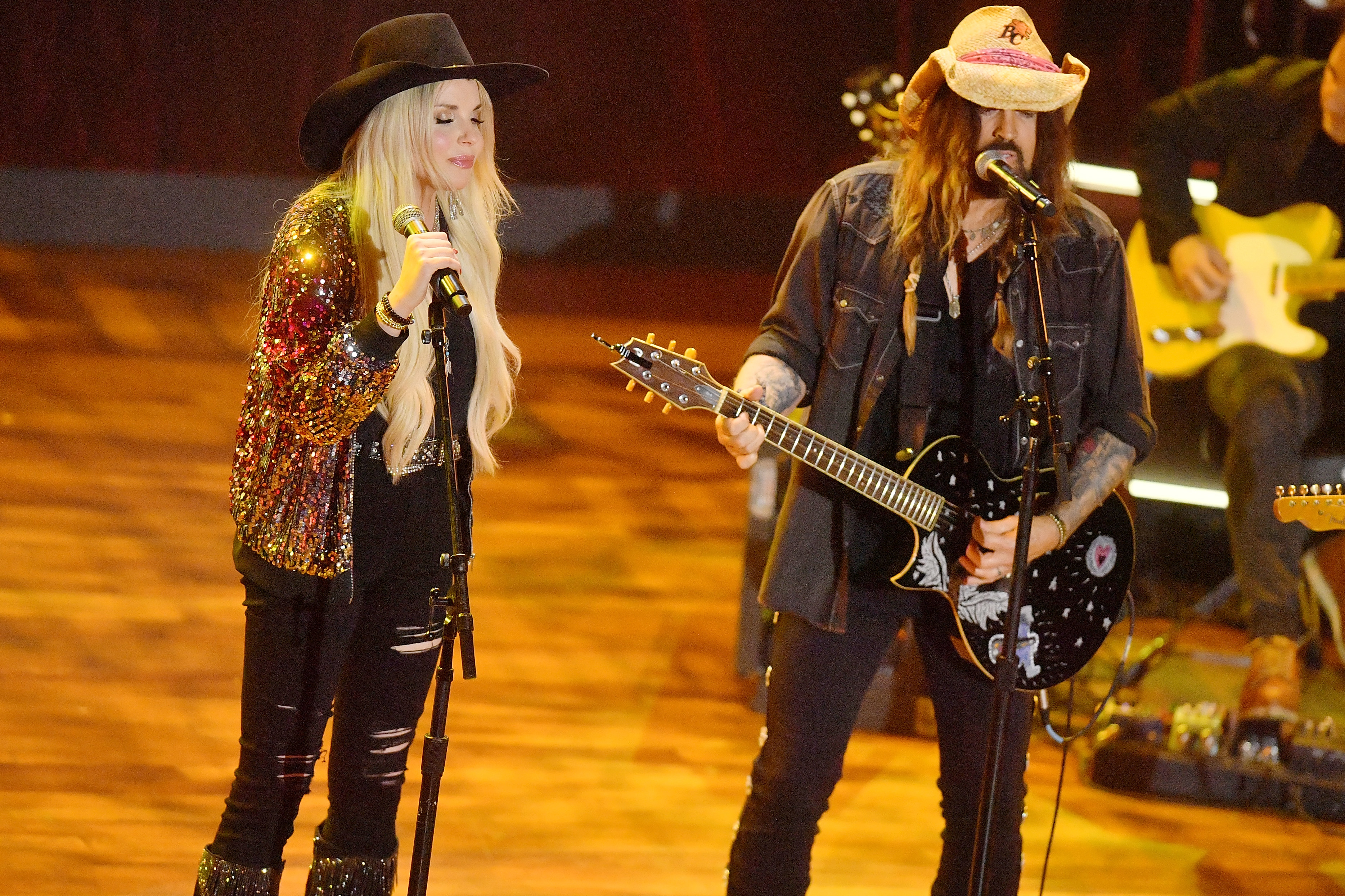 Firerose and Billy Ray Cyrus perform on stage, both wearing hats and casual rock attire with guitars