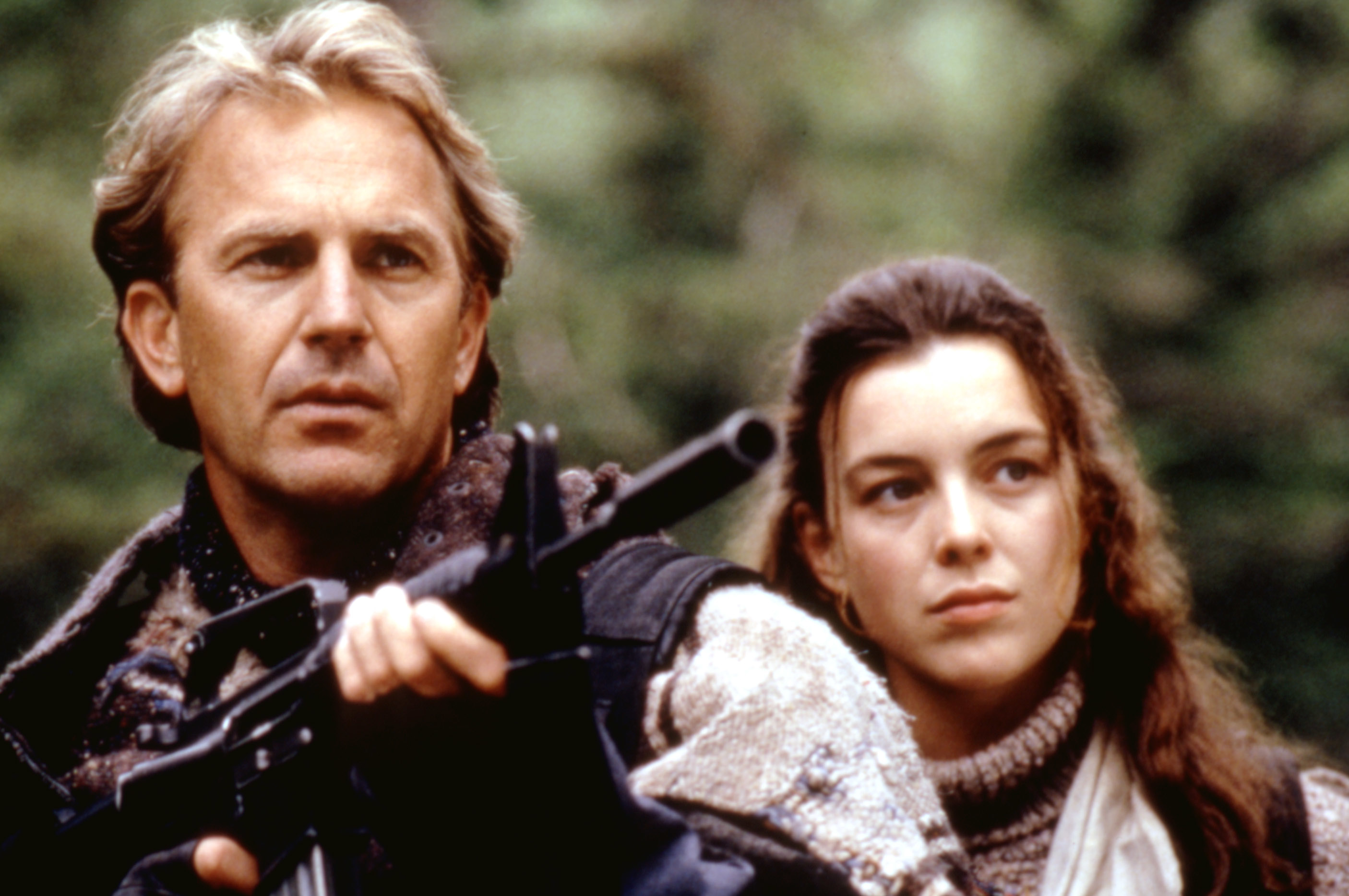Kevin Costner holds a rifle, while Olivia Williams stands beside him, looking in the same direction, in an outdoor setting