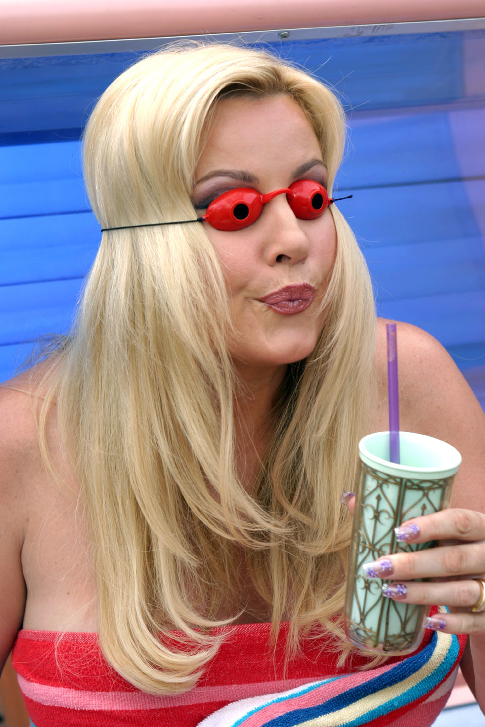 A woman with long blonde hair, wearing small red novelty glasses, holds a decorative cup with a straw