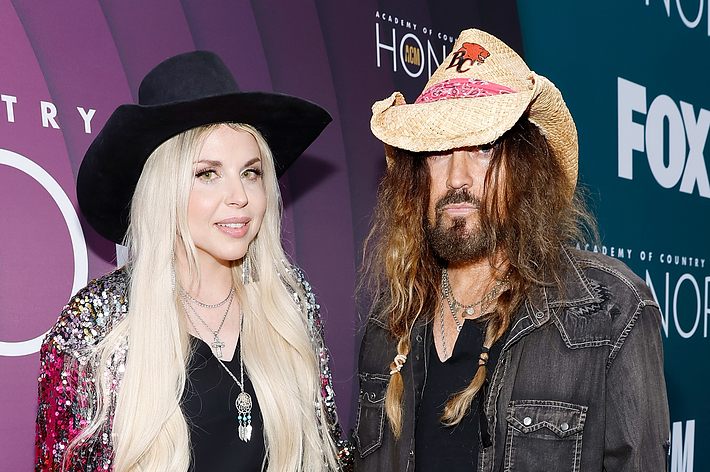 Jessica Caban and Billy Ray Cyrus stand on a red carpet. Jessica wears a sequin jacket and black hat. Billy Ray wears a denim shirt with a cowboy hat