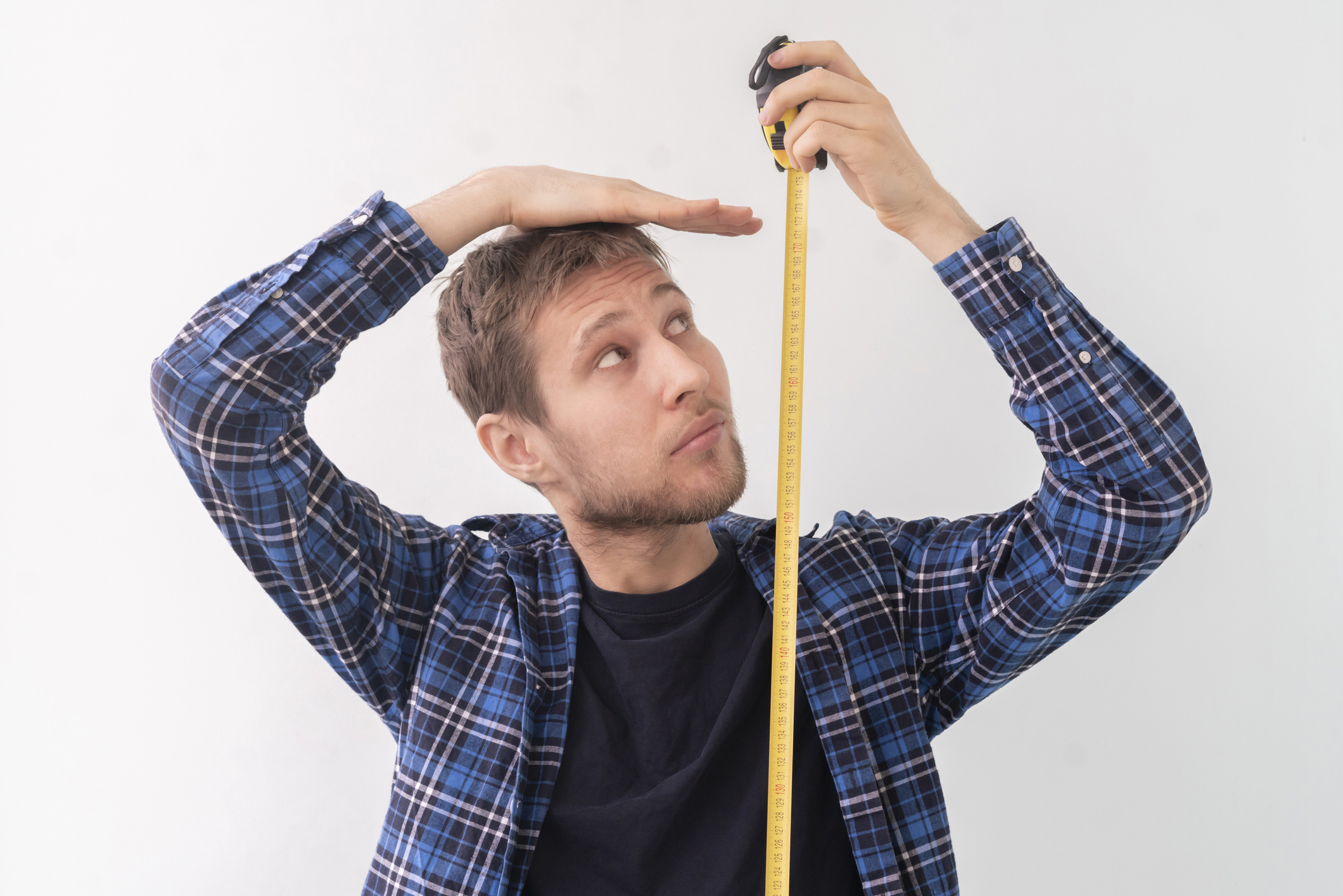 A man in a plaid shirt measuring his height with a measuring tape against a white background