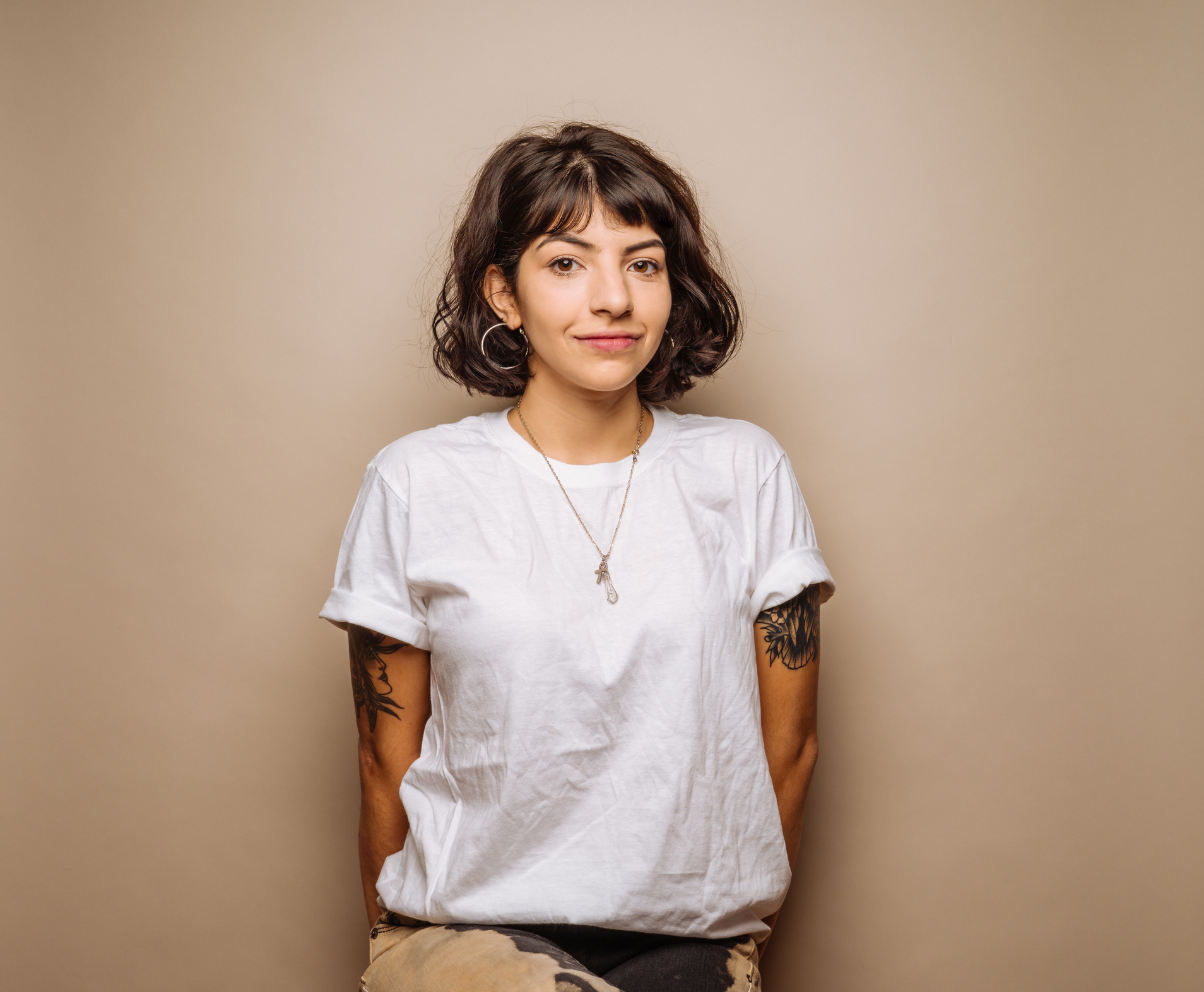 Woman with short wavy hair wearing a white T-shirt and necklaces, standing against a neutral background