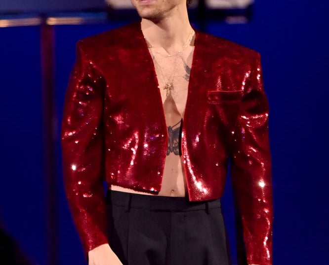 Harry Styles performing, wearing a glittery cropped jacket and black pants, holding a microphone