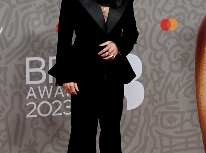 Harry Styles at the BRIT Awards 2023, wearing a black suit with a prominent oversized floral accessory on his shoulder, standing on the red carpet