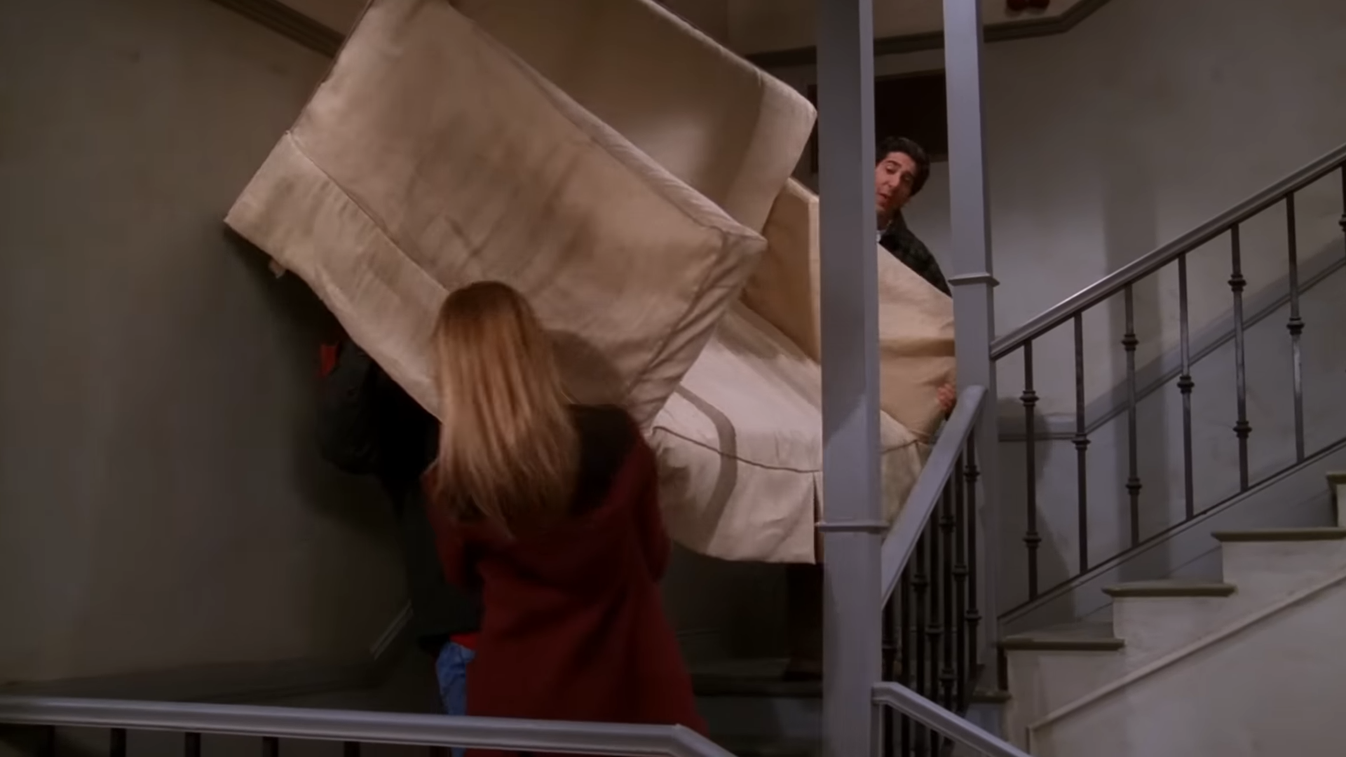 Rachel, Ross, and Chandler struggle to carry a large couch up a narrow staircase in a comical scene from the TV show Friends