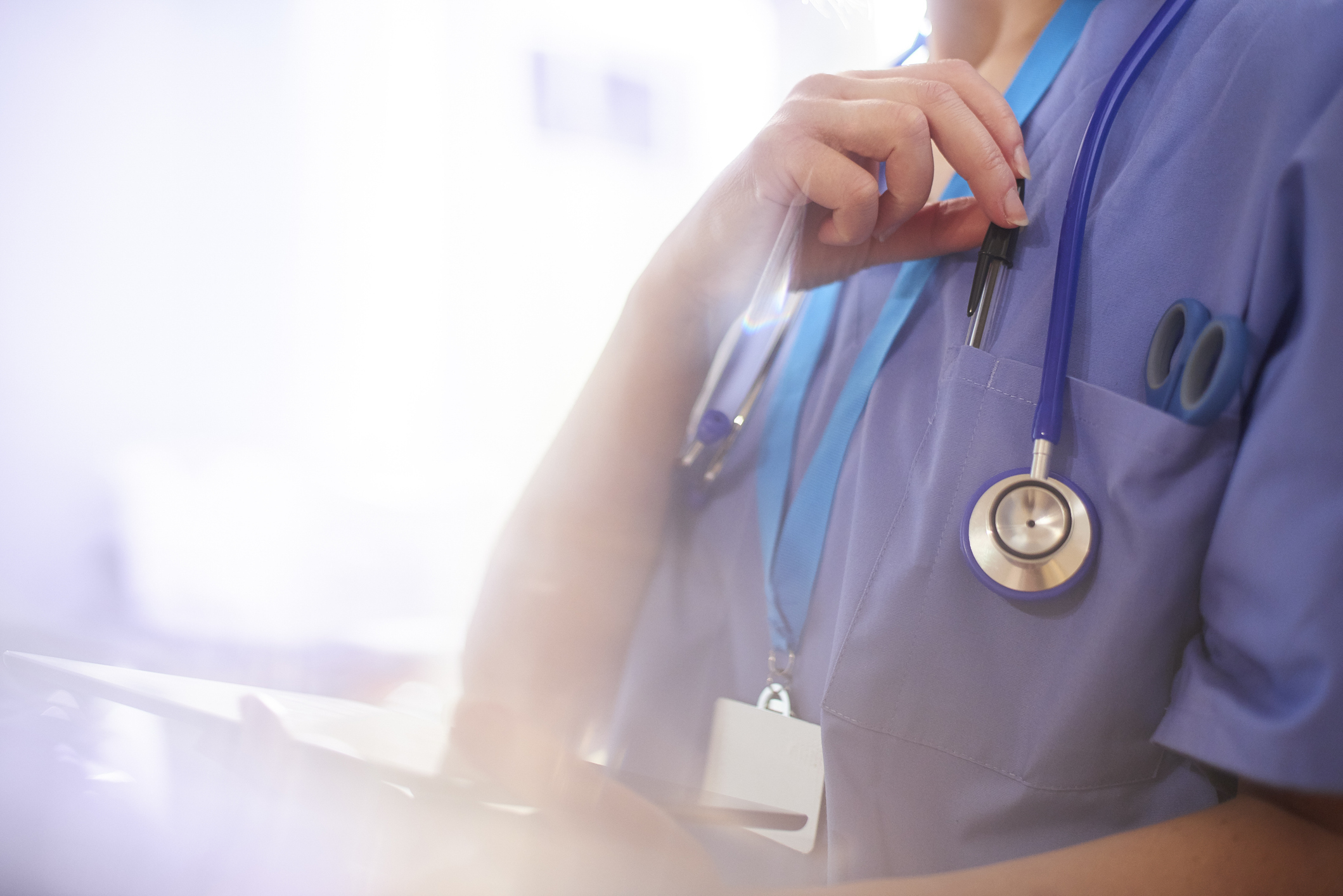 A healthcare worker dressed in scrubs and a stethoscope holds a pen while looking at a clipboard