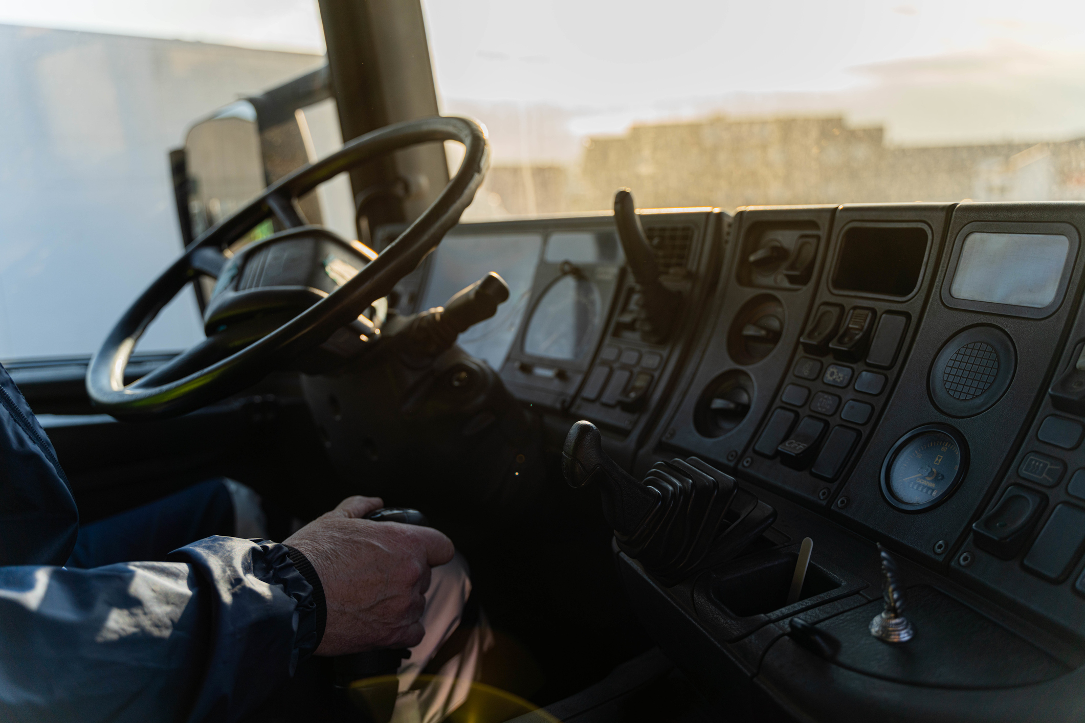 The image shows a person in a driver's seat, holding onto the gear shifter, inside a truck's cockpit with various controls and gauges visible