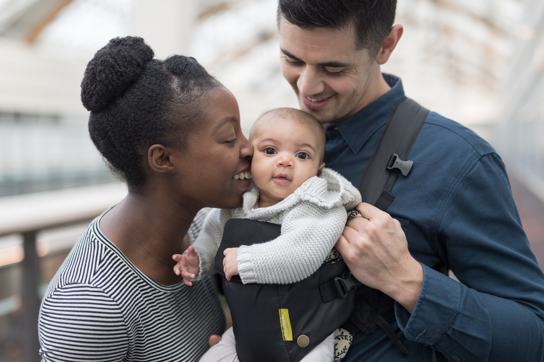 A woman and man hold a baby in a carrier. The woman is smiling and leaning in to kiss the baby while the man looks at the baby with a smile