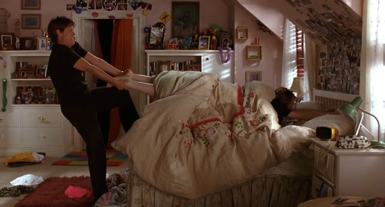 Two people: one is trying to pull the other out of bed in a cluttered bedroom. Photos and decorations cover walls and surfaces