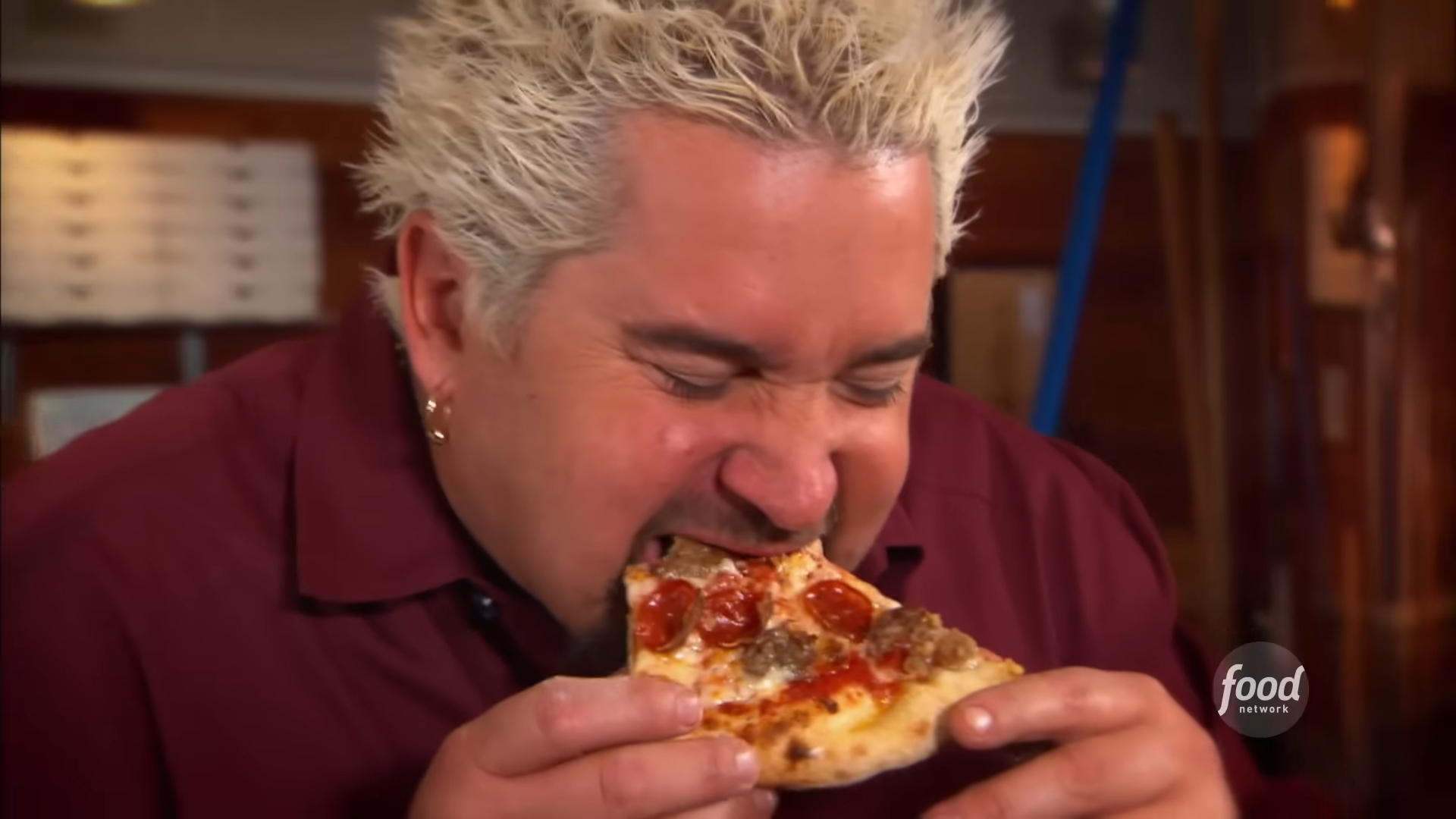 Guy Fieri makan sepotong pizza dengan taburan pepperoni dan sosis.  Logo Food Network ada di pojok kanan bawah