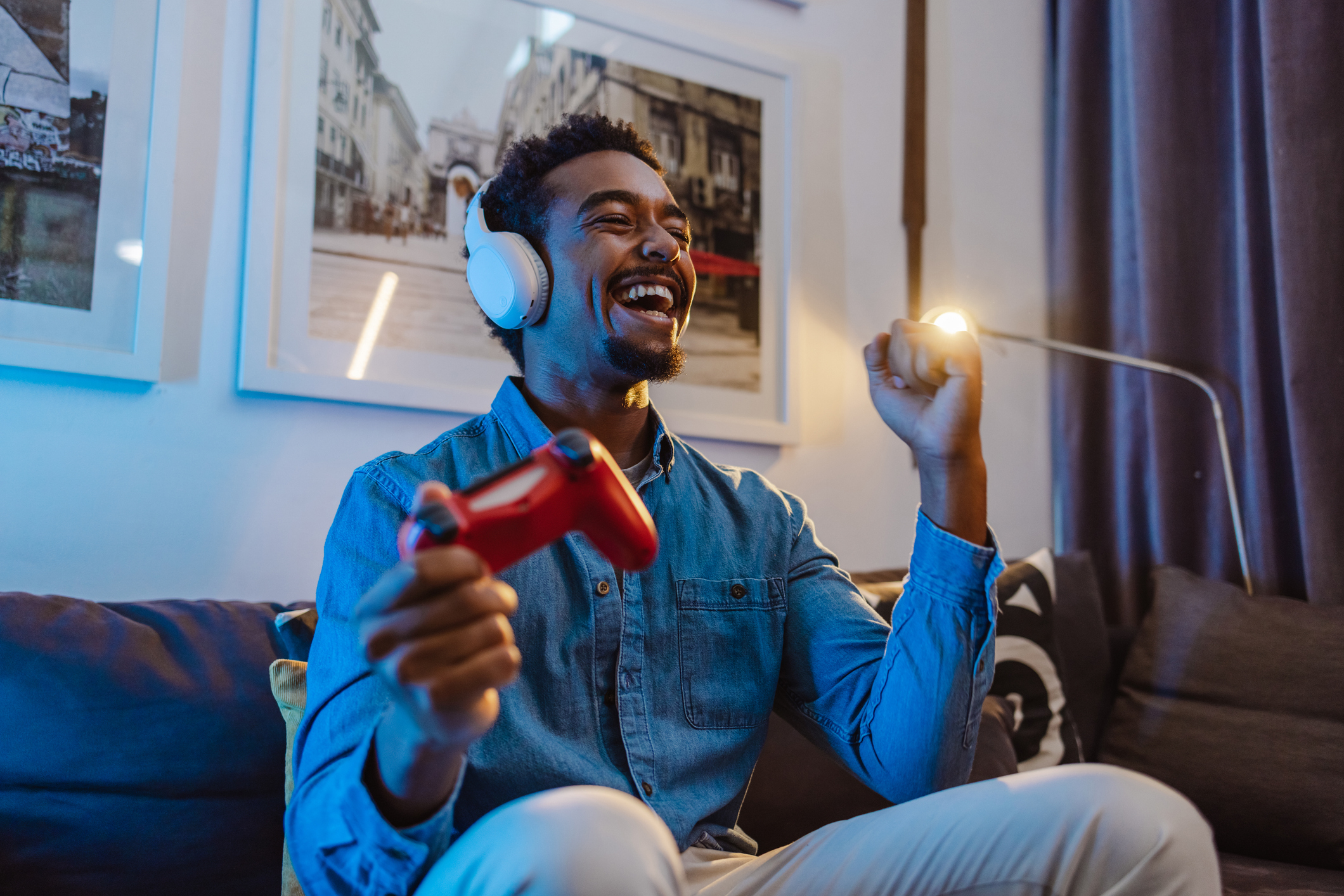 Man wearing headphones, holding a game controller, laughing and cheering while sitting on a couch. Framed photos are on the wall behind him