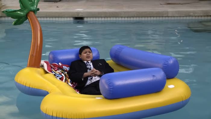 A boy is lying on a yellow inflatable pool float shaped like a car, wearing a suit and tie. There are various items next to him on the float