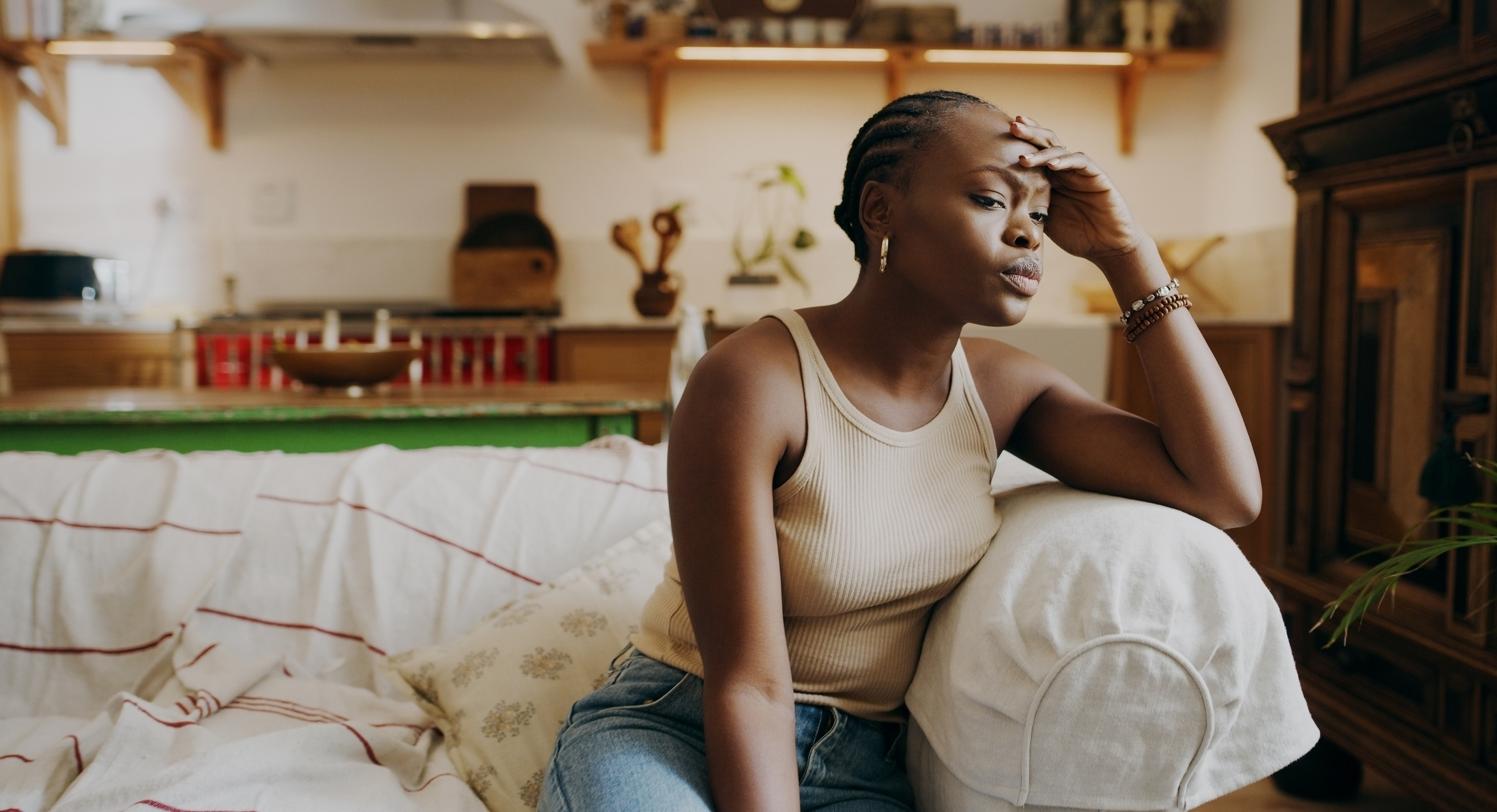 A person sits on a sofa in a cozy kitchen, resting their head on their hand, appearing deep in thought