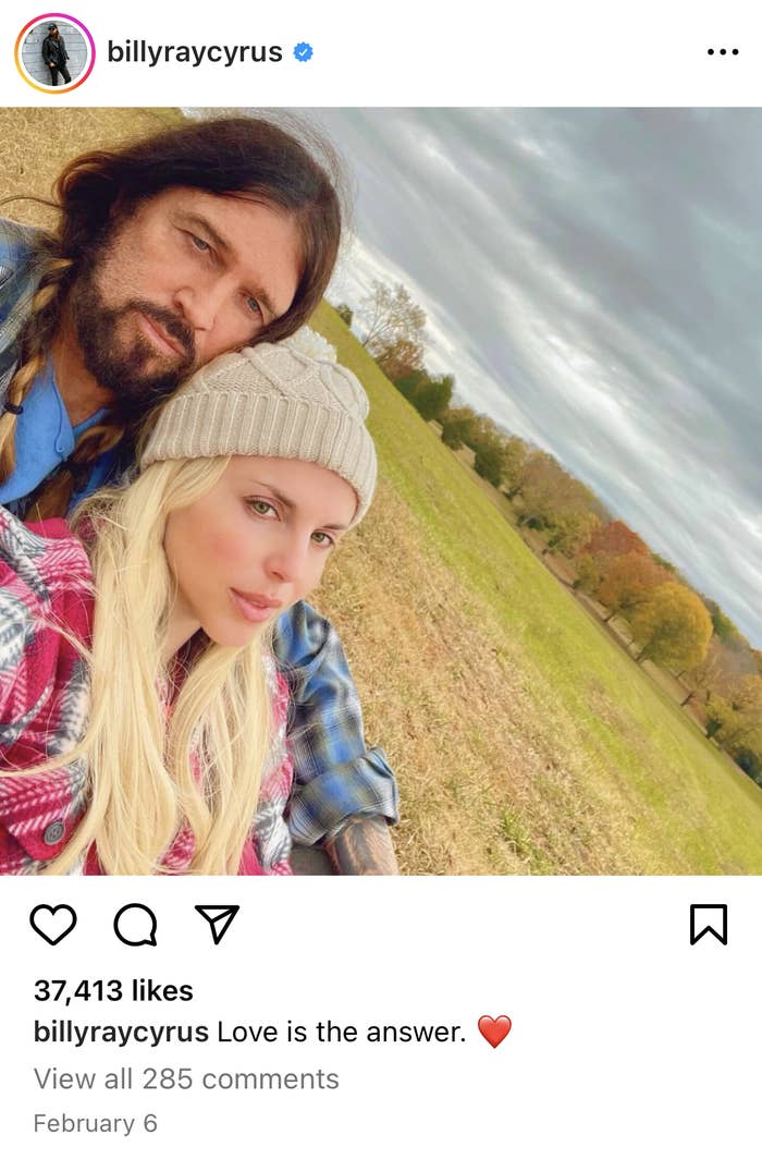 Billy Ray Cyrus and a woman wearing a beanie pose for a selfie outdoors with grass and trees in the background. Caption reads: &quot;Love is the answer. ❤️&quot;