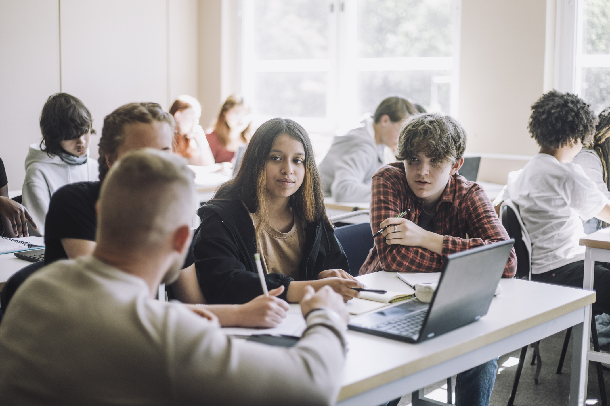 A group of students in a classroom attentively listening to their teacher. They have notebooks and a laptop on their desks