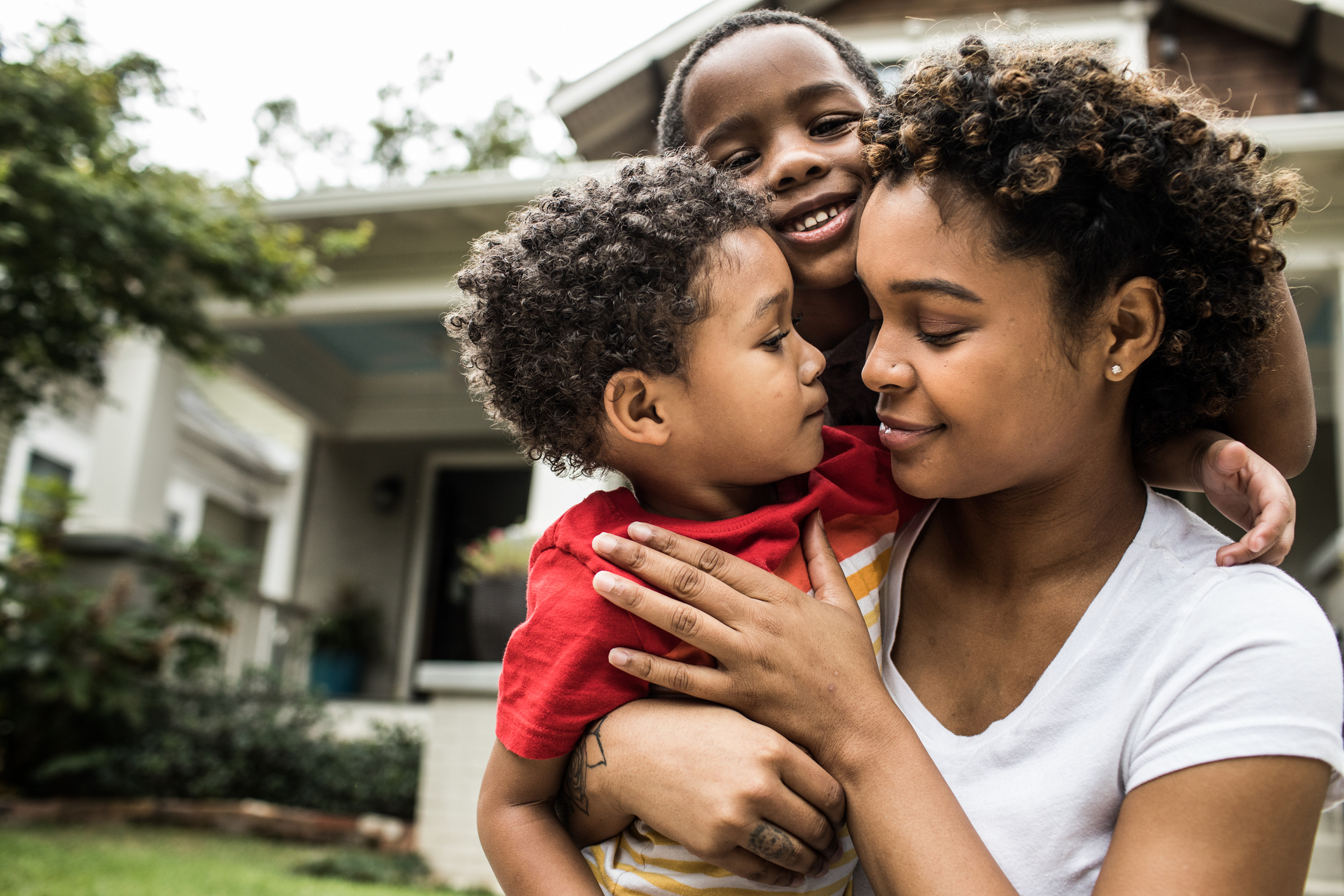 A woman, smiling and holding two children, is gathered closely with them in front of a house