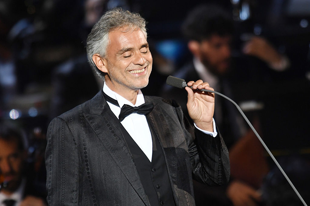 Andrea Bocelli, dressed in a formal black tuxedo, smiles while singing into a microphone during a performance