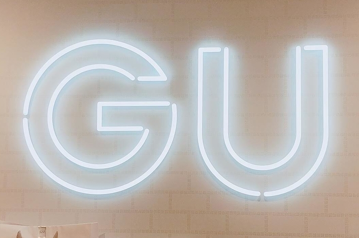GU store wall with a lit GU sign, featuring several white GU branded shopping bags below. Gift wrapping sign visible
