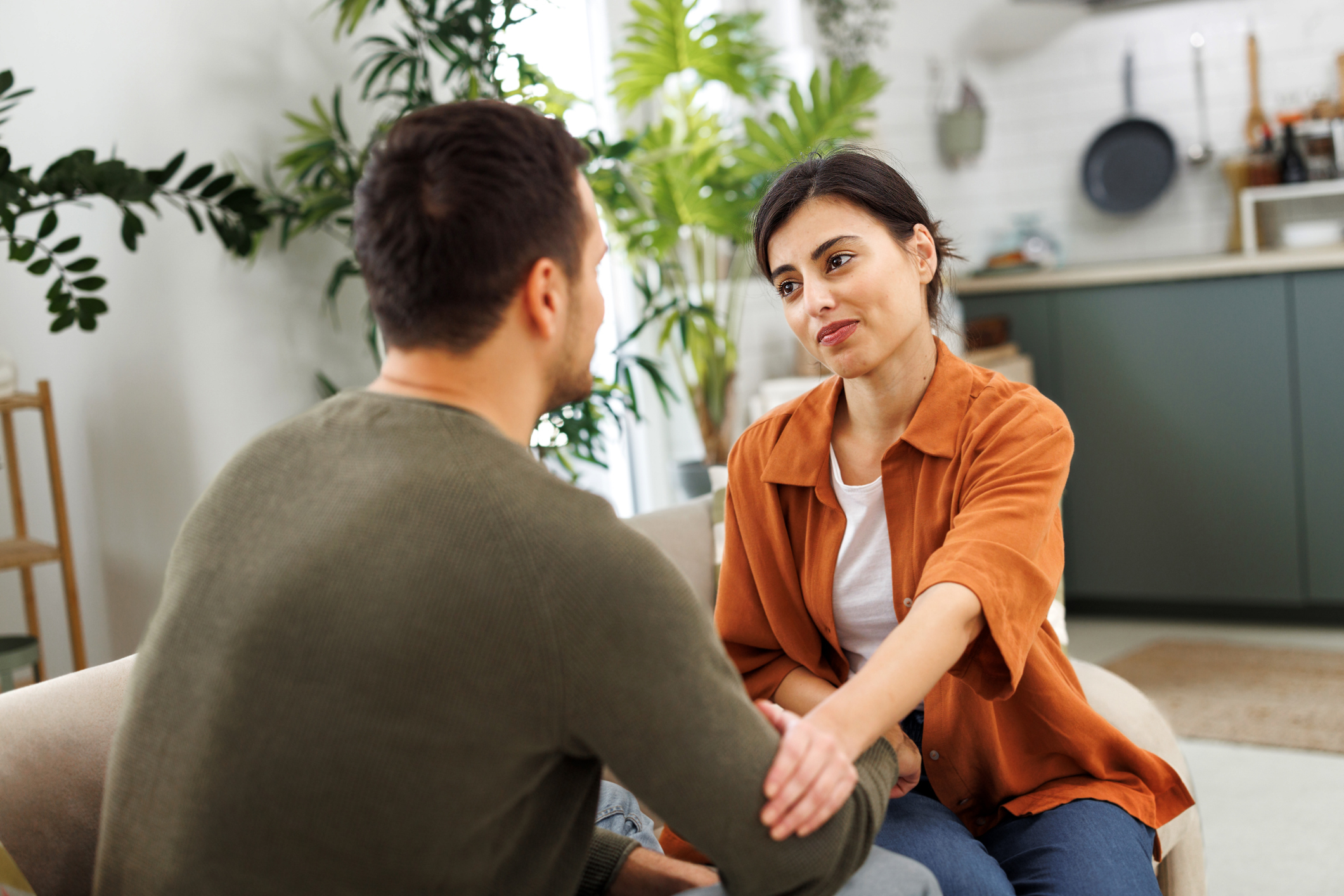 A man and a woman sit on a couch in a modern kitchen. The woman looks concerned and holds the man's hand as he speaks