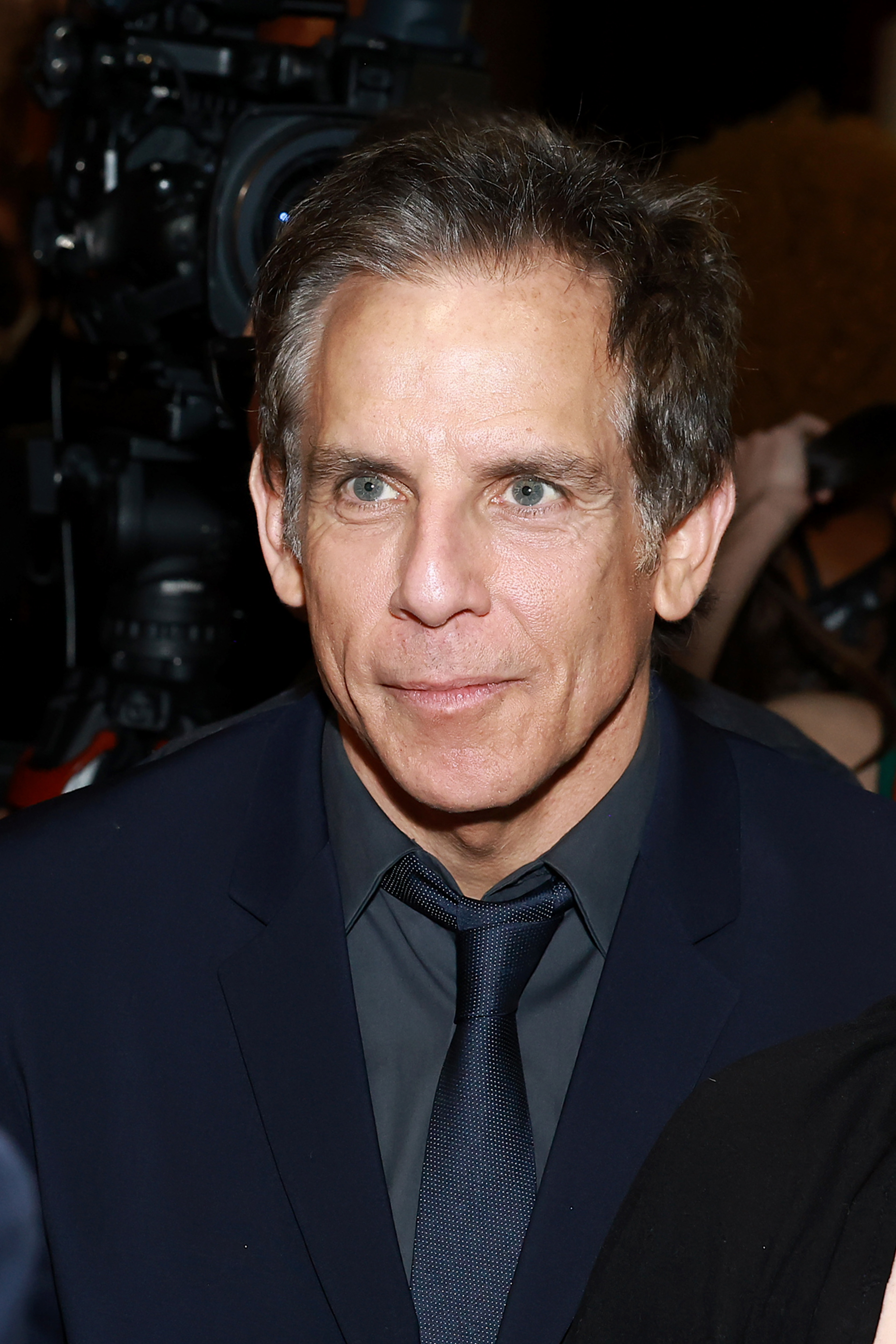 Ben Stiller, wearing a dark suit and tie, attends an event with cameras in the background