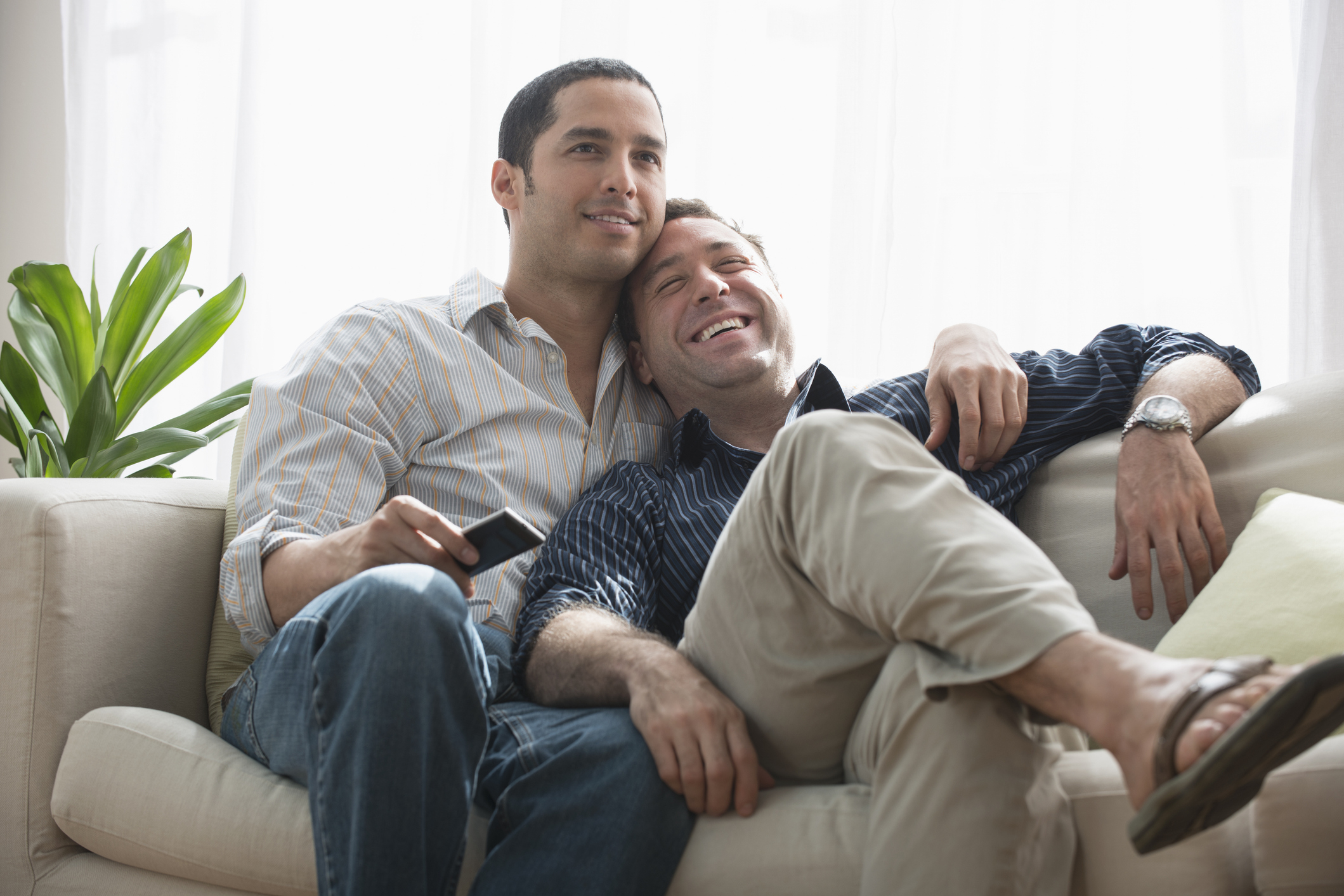 Two men sit closely together on a couch, one leaning on the other's shoulder. They appear content and relaxed, with a remote control in hand
