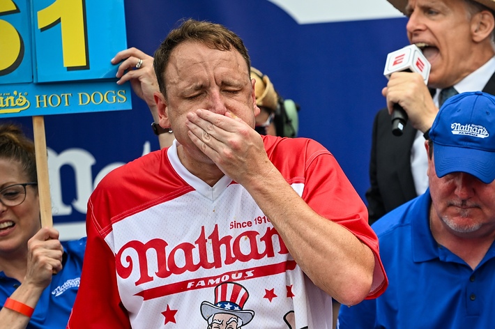 Joey Chestnut, in a Nathan's shirt, competes in a hot dog eating contest, covering his mouth. Announcer and assistants are visible in the background. Sign shows 61