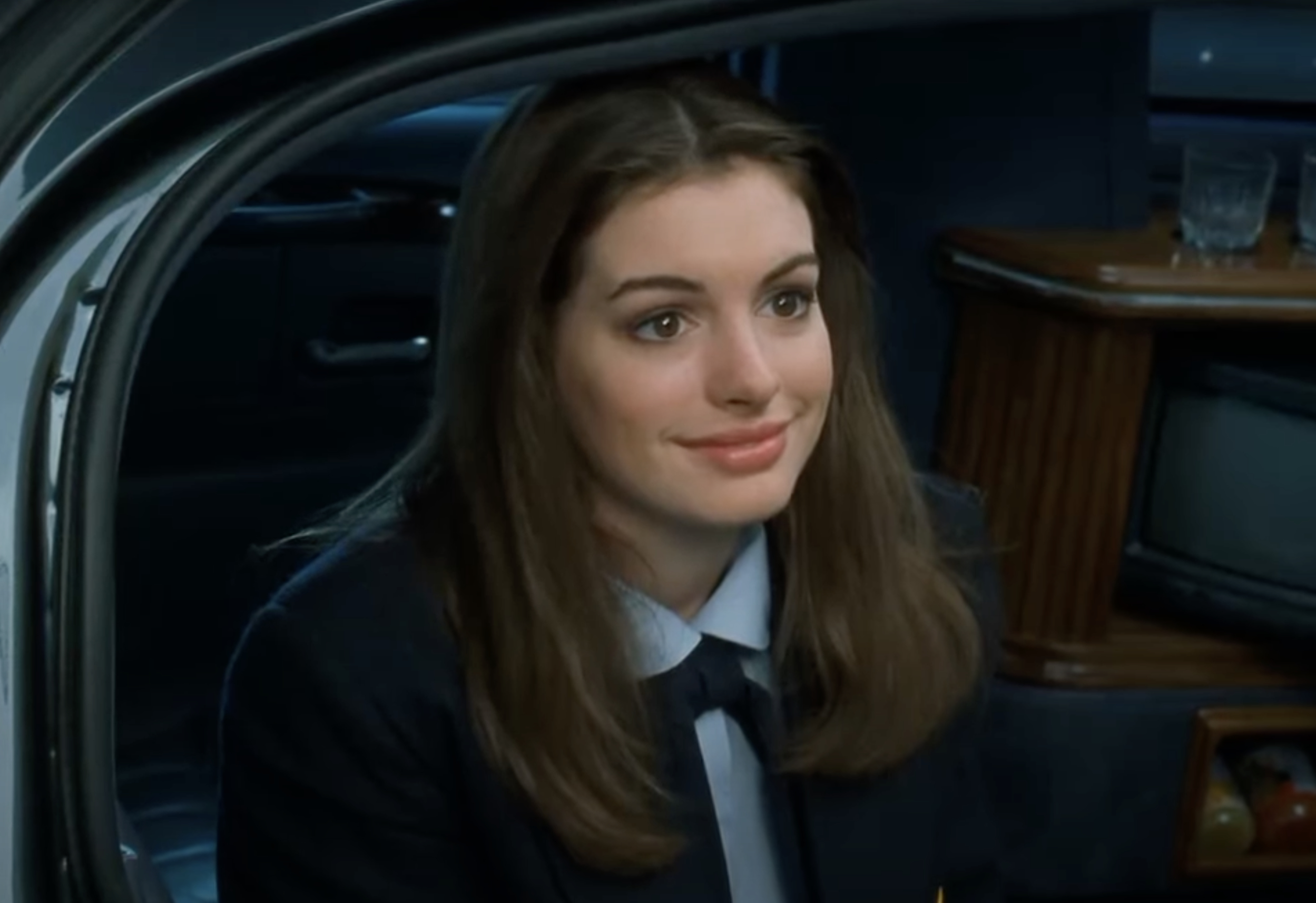 Anne Hathaway, wearing a collared shirt and blazer, smiles while sitting inside a car