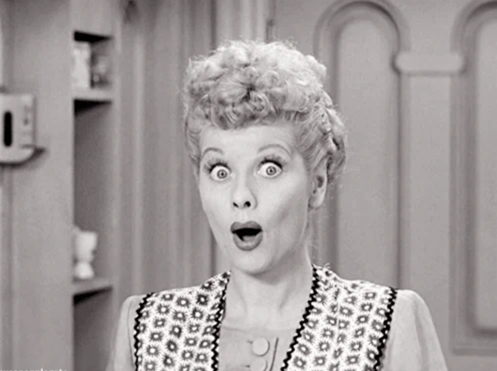 Lucille Ball, with a surprised expression, is dressed in a patterned apron over a light blouse