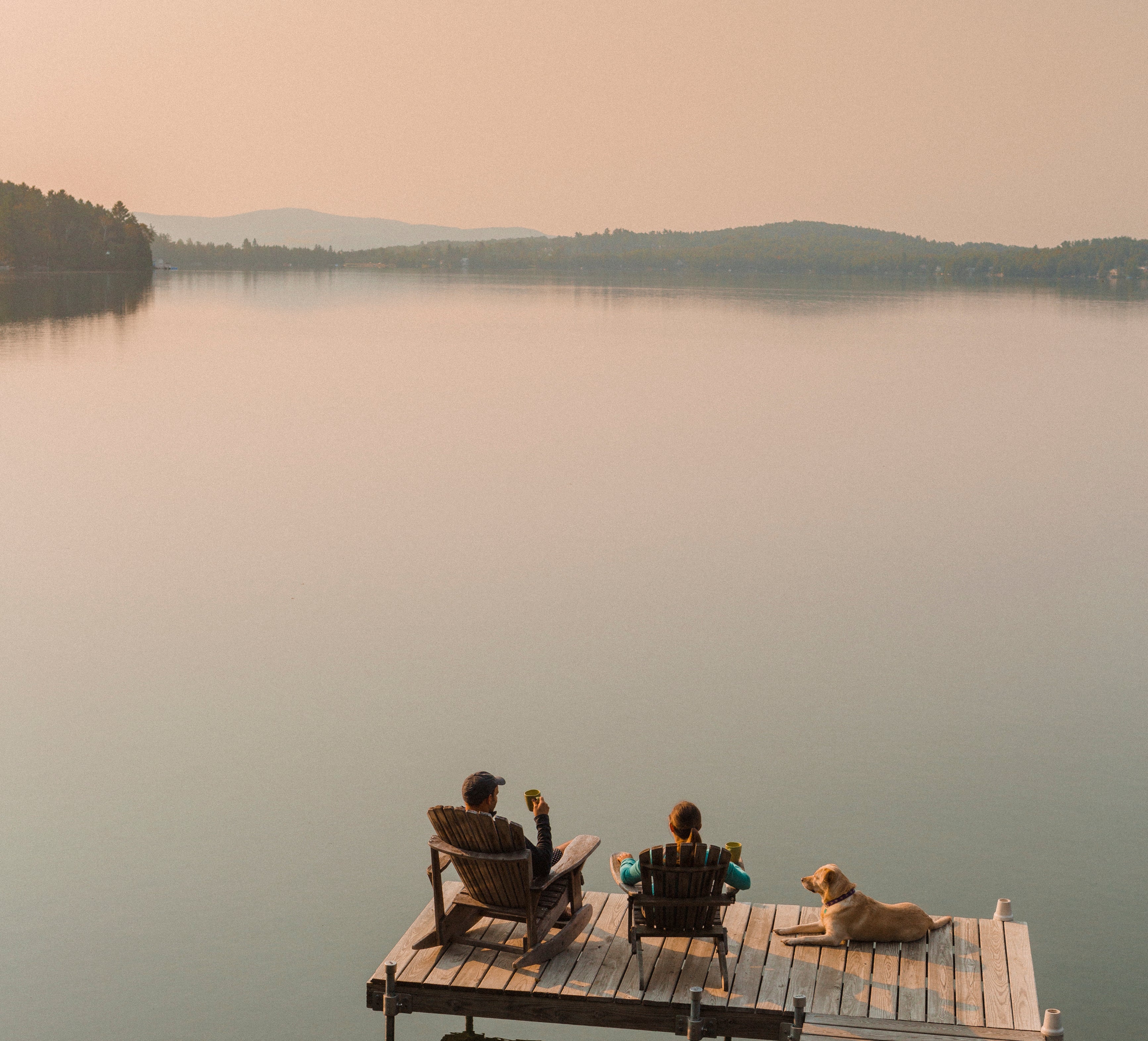 Two people and a dog sit on a dock by a calm lake. The people are sitting on chairs, and the dog is beside them. Mountains are visible in the distance