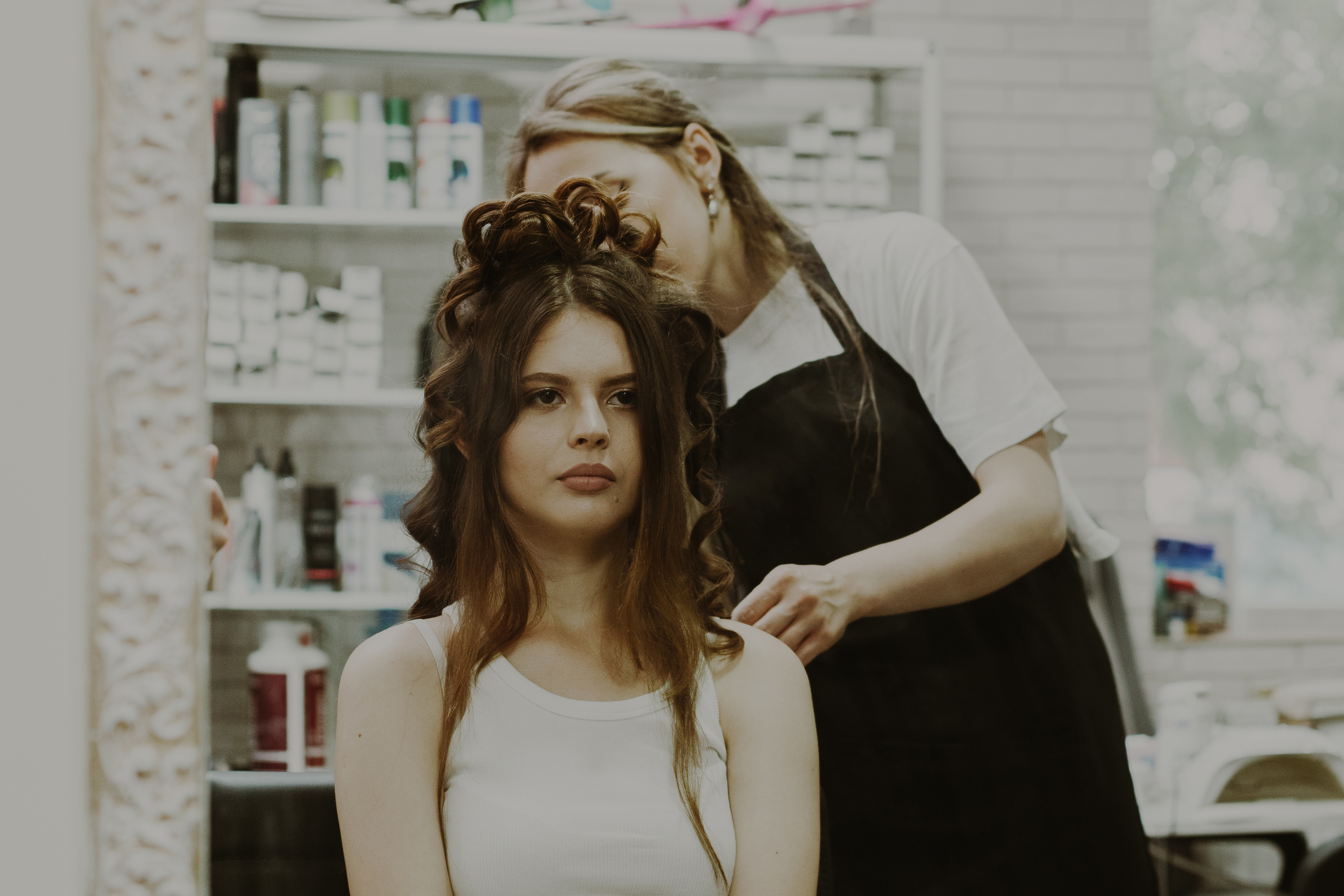 A woman with long hair sits in a salon chair while a stylist works on curling her hair. Shelves with hair products are visible in the background