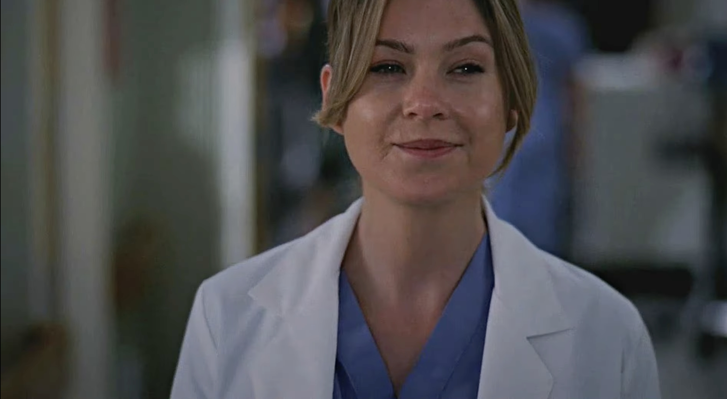 Ellen Pompeo smiles while wearing a light-colored lab coat and blue scrubs in what appears to be a hospital setting