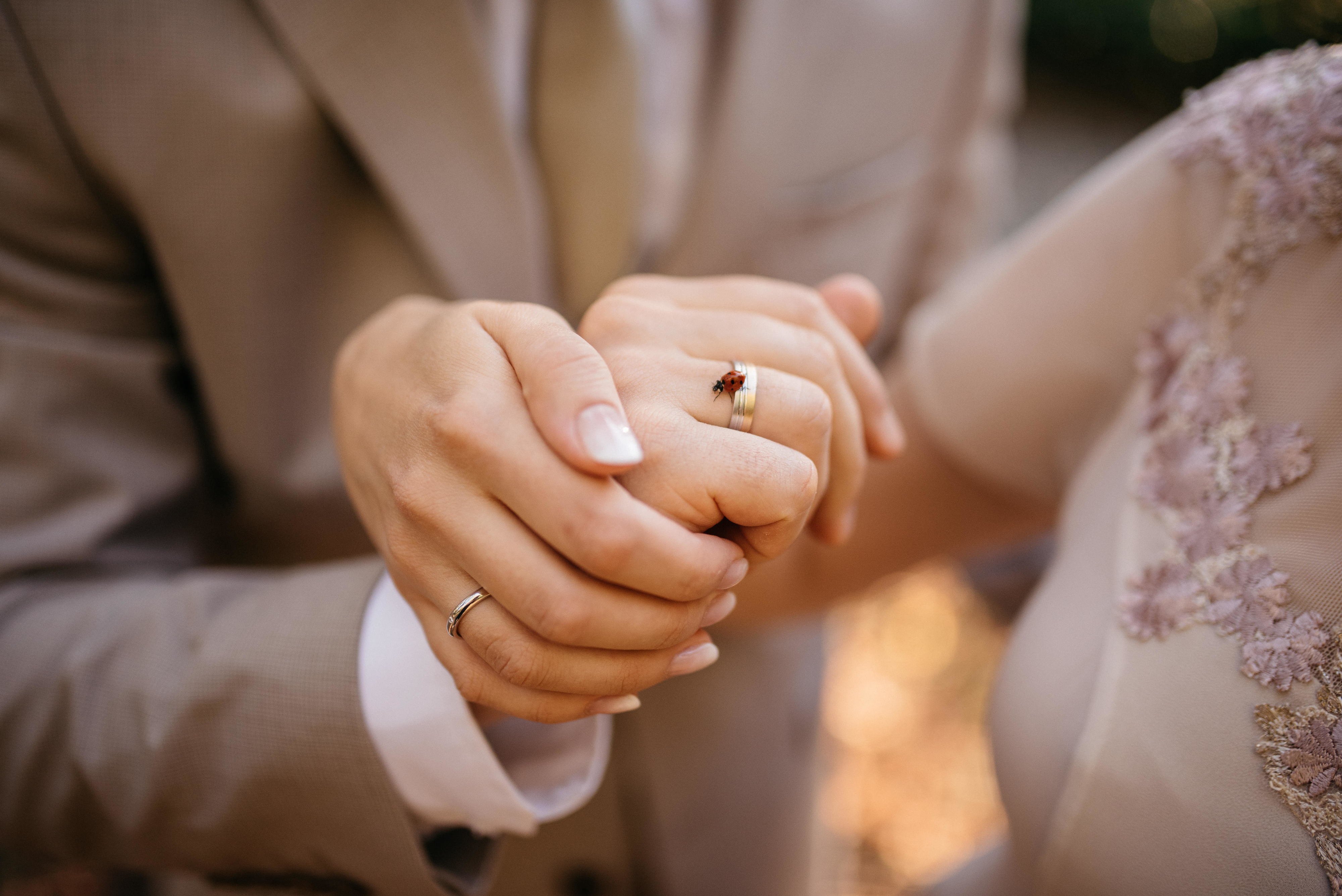 Close-up of a couple holding hands, wearing wedding rings. The individual on the left is in a light-colored suit, and the individual on the right in a floral-embroidered garment