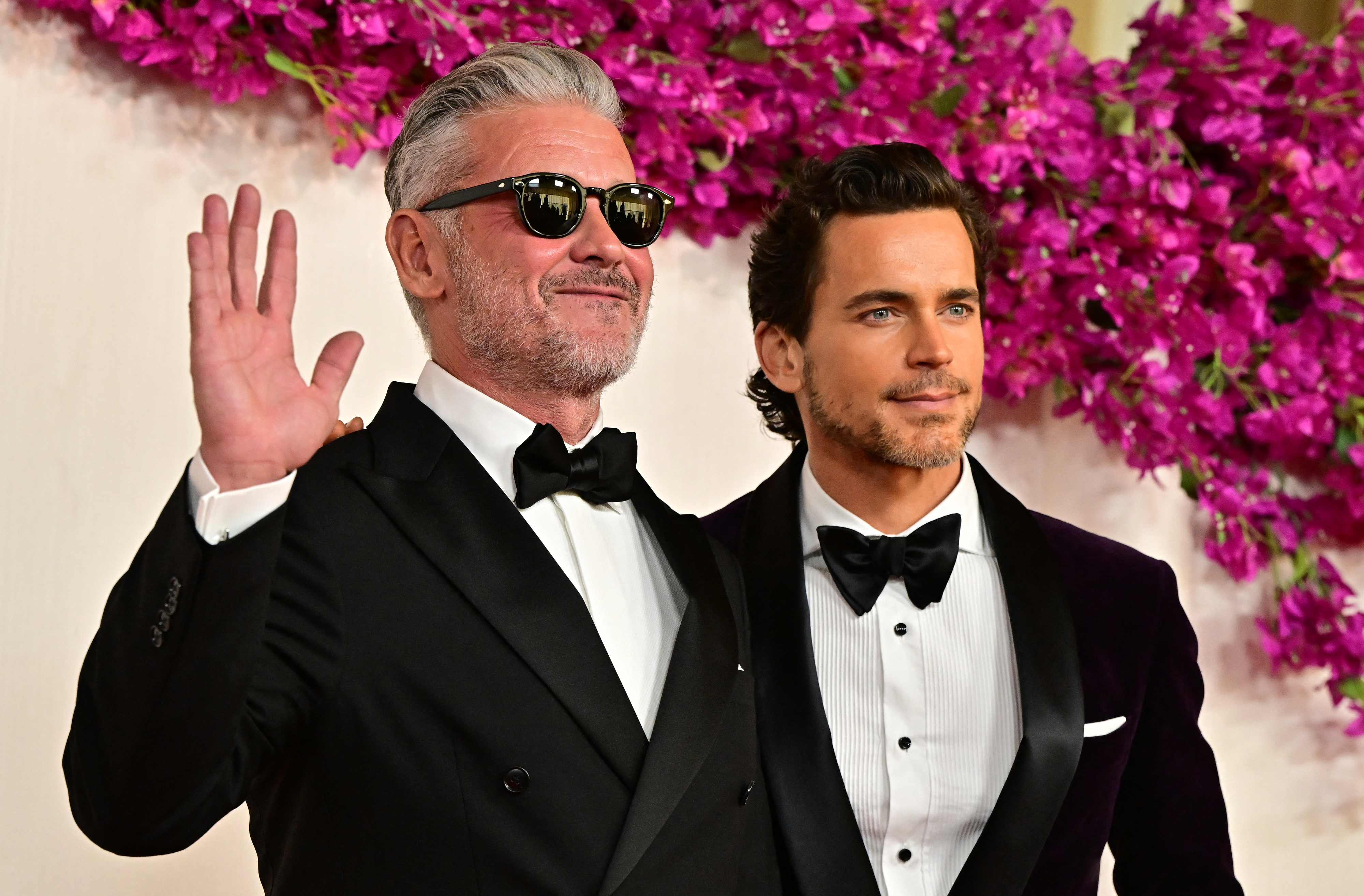 Matt Bomer and Simon Halls on the red carpet wearing tuxedos; Matt with a bow tie and Simon with sunglasses, waving, against a floral backdrop