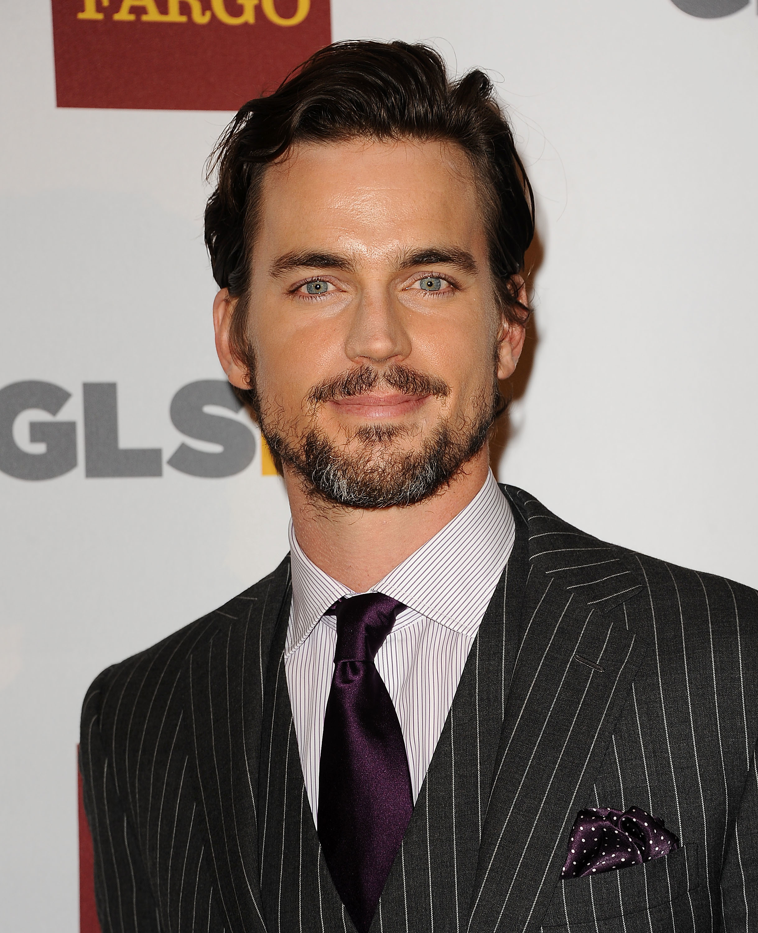 Matt Bomer at an event, wearing a pinstriped suit, striped shirt, and dark silk tie with a matching pocket square, smiling and looking at the camera