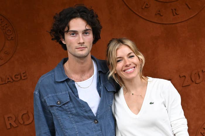 Sienna Miller and Oli Green pose together. Sienna wears a white top, and Oli wears a blue button-up shirt over a white t-shirt