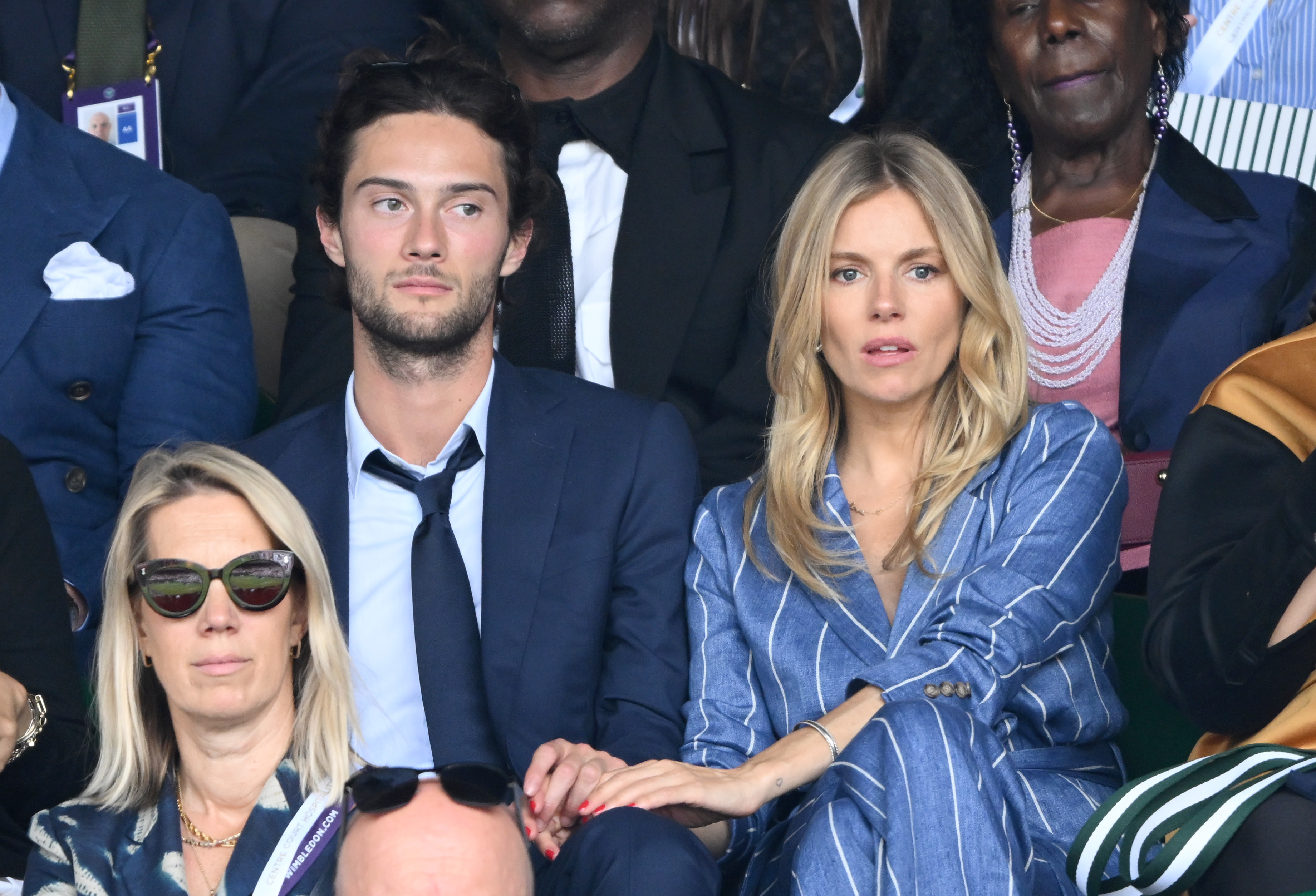 Sienna Miller, in a pinstriped suit, sits with Oli Green at a formal event. They hold hands, looking attentive. Others are also seated around them