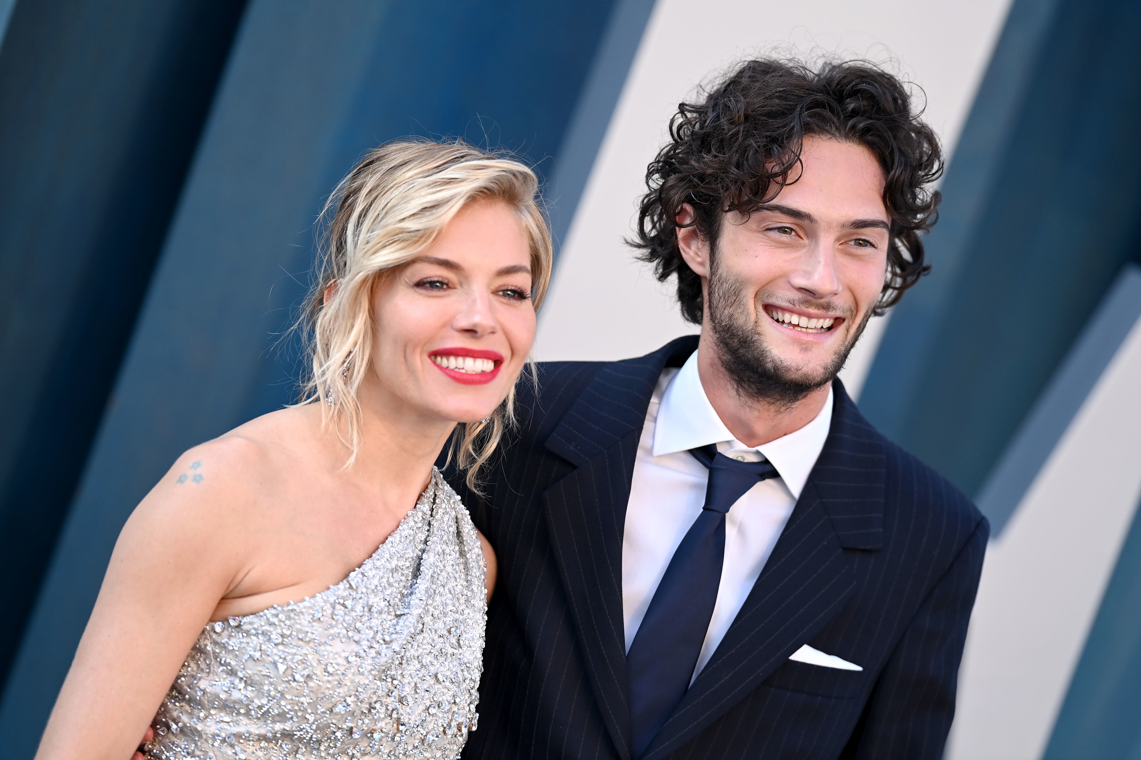 Smiling Sienna Miller in a sparkling one-shoulder dress and Oli Green in a suit and tie, posing together at a formal event