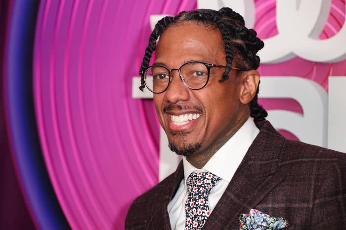 Nick Cannon smiles while wearing a dark blazer, floral tie, glasses, and braided hairstyle at a public event with a vibrant background