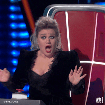 Kelly Clarkson on The Voice, expressing surprise with her mouth open and hands up, seated at her judge's chair