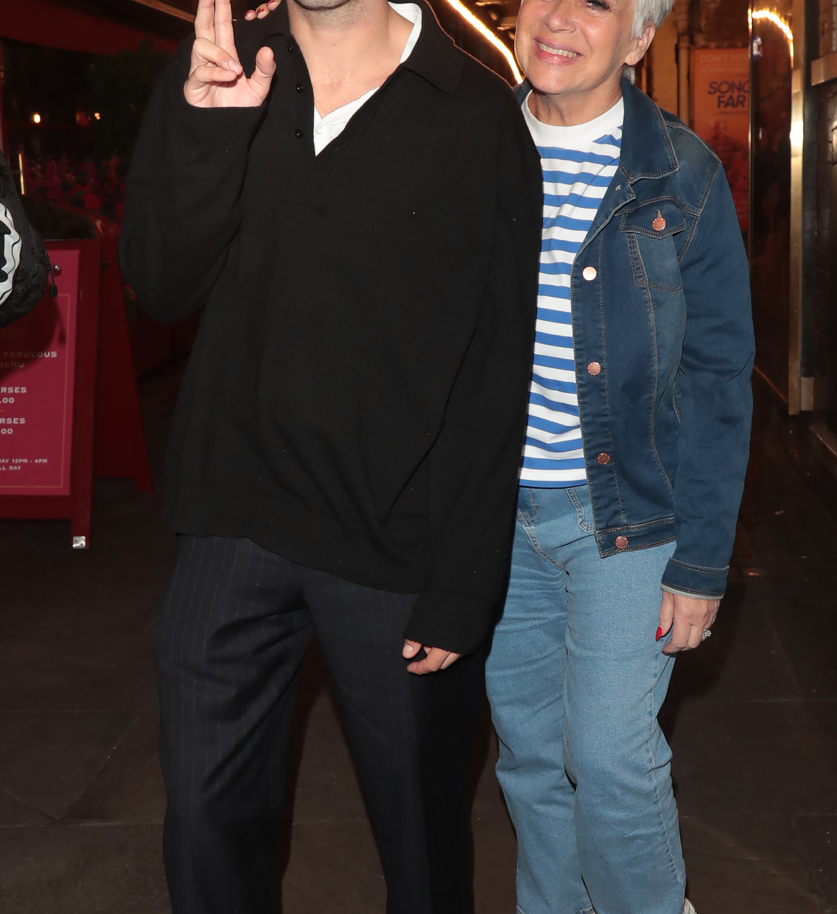 Matty Healy and Denise Welch smile and pose on a street wearing casual outfits with Denise in a denim jacket and striped shirt and Matty in a dark shirt and pants