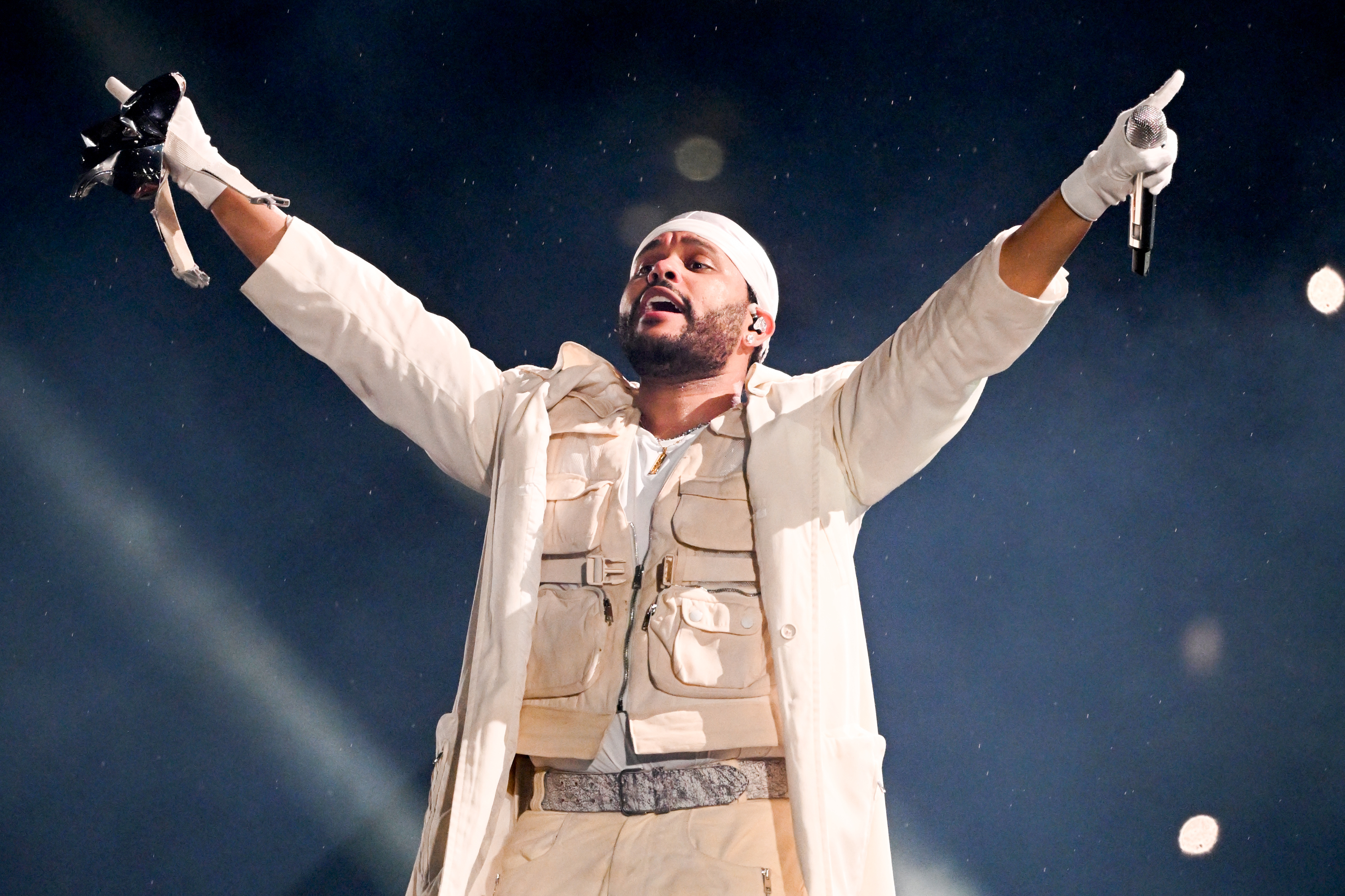 The Weeknd performing on stage, wearing a white outfit with utility vest and gloves, holding a microphone with arms raised