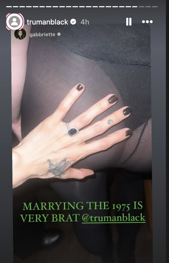 A hand with dark nail polish and tattoos, wearing a ring, is shown against a sheer fabric. Text overlay says, &quot;MARRYING THE 1975 IS VERY BRAT @trumanblack @gab.briette&quot;