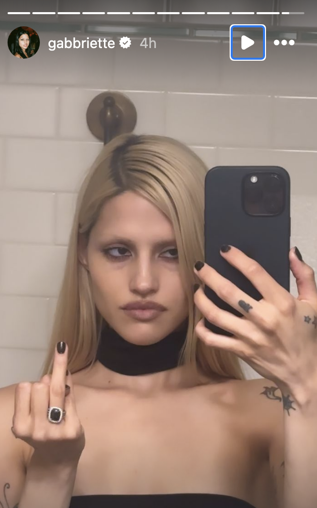 Gabbriette takes a mirror selfie, holding her phone in one hand and showing her middle finger with the other. She has long blonde hair and wears a choker necklace