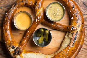 Large pretzel on a wooden serving board with three dipping sauces: cheese, mustard, and a small bowl of pickles
