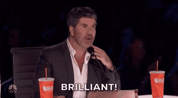Simon Cowell wearing a suit and sitting at a table with two drink cups, gesturing and saying "Brilliant!" on America's Got Talent